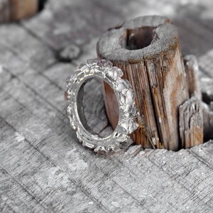 Puede incluir: Un anillo de plata con un dise&ntilde;o texturizado y peque&ntilde;as piedras rosas. El anillo est&aacute; sobre un trozo de madera desgastada.
