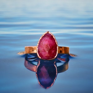 May include: A rose-coloured gemstone ring with a pear-shaped stone set in a gold band. The ring is floating on the surface of blue water.