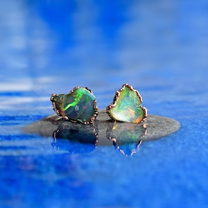 以下が含まれることがあります： 虹色の緑色のオパール石がついた、銅線で巻かれたイヤリングのペア。イヤリングは、青い背景の灰色の石の表面の上に置かれています。