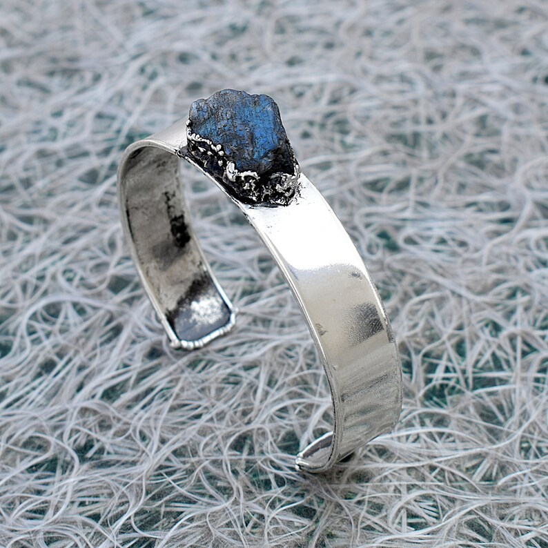 May include: A silver cuff bracelet featuring a blue labradorite gemstone set in a silver bezel. The bracelet is open and has a smooth, polished finish.