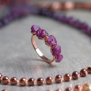 May include: A rose gold ring featuring a band of small, raw ruby stones. The stones are a deep pink colour and have a natural, unpolished look.