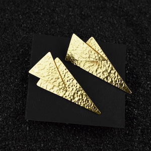 May include: A pair of gold-toned earrings with a geometric triangle design. The earrings are made of hammered metal and have a textured surface.