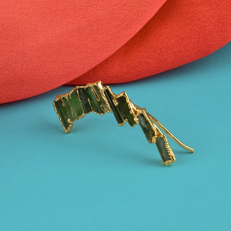 Puede incluir: Pendiente de oro con un dise&ntilde;o curvo adornado con m&uacute;ltiples piedras preciosas verdes.