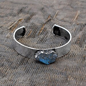 May include: A silver cuff bracelet featuring a large, blue-green gemstone set in the centre. The gemstone has a rough, natural appearance.
