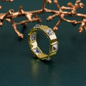 Puede incluir: Un anillo de oro con piedras cuadradas transparentes engastadas en la banda. El anillo está sobre un fondo verde.