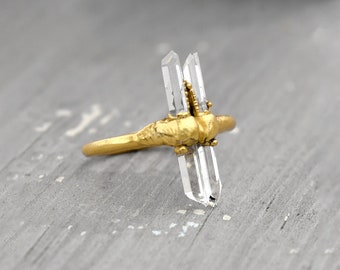 Könnte beinhalten: Ein goldener Ring mit zwei klaren Quarzkristallen, die in der Mitte eingesetzt sind. Der Ring ist aus strukturiertem Metall gefertigt und hat ein rustikales Aussehen.