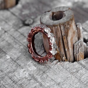 Puede incluir: Un anillo de oro rosa con una banda de piedras facetadas transparentes engastadas en un metal texturizado. El anillo descansa sobre un trozo de madera desgastada.