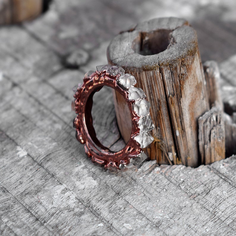 Puede incluir: Un anillo de oro rosa con una banda de piedras facetadas transparentes engastadas en un metal texturizado. El anillo descansa sobre un trozo de madera desgastada.