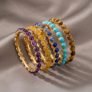 Può includere: Un set di cinque anelli color oro, ciascuno caratterizzato da una fascia continua di piccole perle di pietre preziose rotonde e levigate. Gli anelli presentano colori distinti tra cui ametista viola, citrino giallo, lapislazzuli blu, turchese e occhio di tigre marrone.