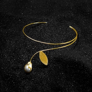Puede incluir: Un brazalete dorado con un diseño floral. El brazalete presenta una banda delgada y curvada. Un tallo se extiende desde la banda, sosteniendo una hoja dorada y una perla nacarada en forma de lágrima. Fondo negro.
