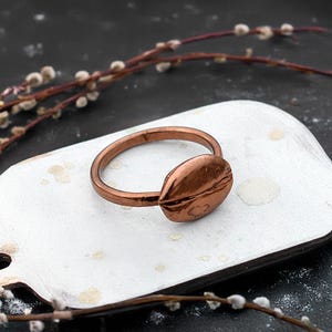 May include: A copper ring with a textured oval-shaped band. The ring is on a white surface.