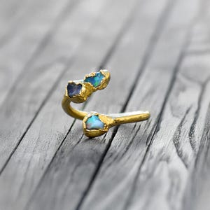 May include: A gold ring with three raw gemstones. The ring is made of gold and has a unique, organic shape. The gemstones are a blue sapphire and two opals.