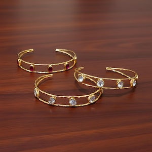 May include: Three gold-colored open cuff bracelets. One bracelet has oval-shaped red gemstones, and the other two have clear gemstones. The bracelets are displayed on a dark brown wooden surface.