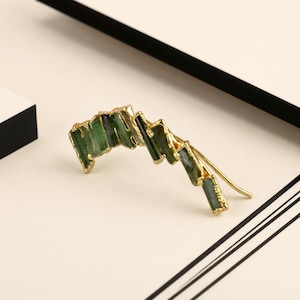 May include: An ear climber earring featuring a series of raw, rectangular green tourmaline crystals set in a gold-toned metal frame. The crystals are arranged in a curved, ascending line, with a thin gold wire post extending from the back.