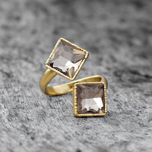 May include: A gold ring with two square-shaped clear stones. The stones are set in a gold frame with a decorative edge.