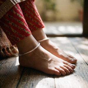 May include: A pair of simple, smooth, polished silver anklets worn around the ankles. The anklets are thin, circular, and metallic in appearance, contrasting with the red and yellow striped fabric of the trousers visible above the feet.
