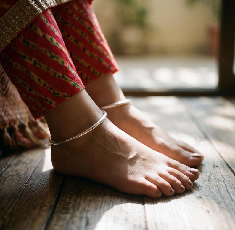 May include: A pair of simple, smooth, polished silver anklets worn around the ankles. The anklets are thin, circular, and metallic in appearance, contrasting with the red and yellow striped fabric of the trousers visible above the feet.