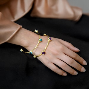 Puede incluir: Dos pulseras de oro con piedras preciosas turquesa y amatista. Las pulseras están apiladas en la muñeca de una persona.