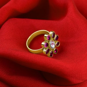 May include: A gold ring with a flower-shaped design. The center of the flower has a clear stone surrounded by eight smaller pink stones.