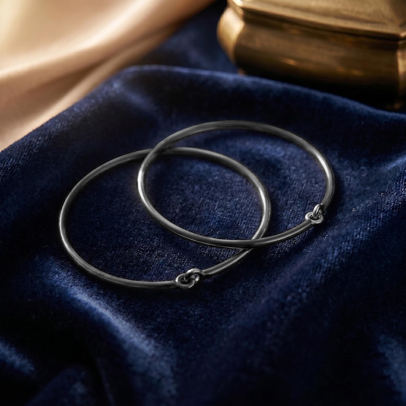 May include: Two thin, dark gray metal bangle bracelets with a smooth, circular design. Each bracelet features a small, integrated hook-and-eye style clasp mechanism that allows the circular band to connect securely at a single point.
