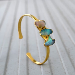 Op de afbeelding: Een gouden armband met drie opaalstenen. De stenen zijn lichtblauw en roze.