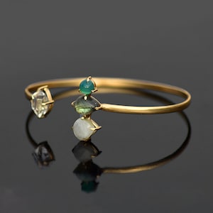 May include: A gold bangle bracelet featuring three gemstones. The top gemstone is green, the middle gemstone is green and grey, and the bottom gemstone is white.