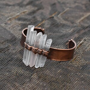 May include: A copper cuff bracelet with clear quartz crystals set in a row across the top. The bracelet is on a brown textured surface.