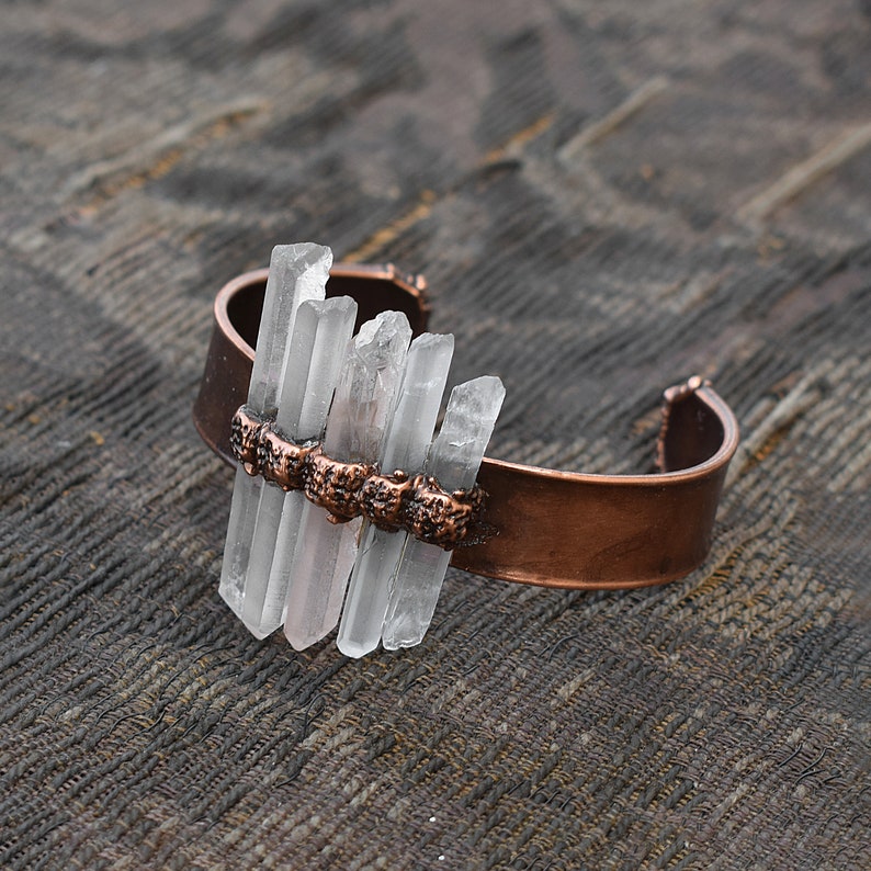 May include: A copper cuff bracelet with clear quartz crystals set in a row across the top. The bracelet is on a brown textured surface.