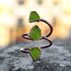 Op de afbeelding: Een koperen ring met drie groene glazen stenen die in het metaal zijn gezet. De ring is spiraalvormig gekruld.