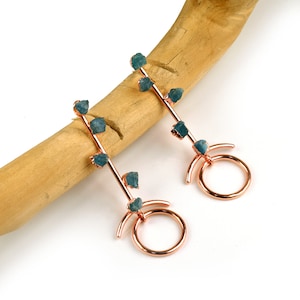 May include: A pair of rose gold earrings with a long, thin bar adorned with multiple blue gemstone accents. Each earring features a circular hoop at the bottom.