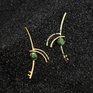 Puede incluir: Un par de pendientes de oro con una piedra preciosa verde en el centro de cada pendiente. Los pendientes están hechos de metal fino y curvado y tienen un diseño delicado y moderno.