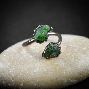 May include: A silver ring with two green gemstones set in the metal. The ring is open and adjustable.