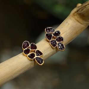Op de afbeelding: Een paar gouden oorbellen met een cluster van donkerrode edelstenen. De oorbellen zijn gevormd als een bloem met vijf bloemblaadjes.