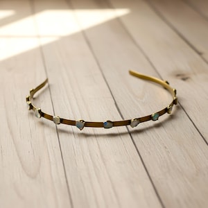 May include: A gold-coloured headband featuring small, irregularly shaped, light-coloured stones evenly spaced along the front. The headband rests on a light-coloured wooden surface, with soft lighting.