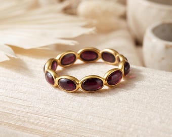 Può includere: Un anello color oro caratterizzato da una fascia continua di pietre preziose granato rosso scuro di forma ovale, incastonate in una montatura di metallo liscio. L'anello presenta una serie di pietre lucide disposte in cerchio.