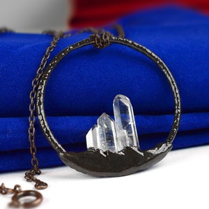 Op de afbeelding: Een zilveren ketting met een zwarte metalen cirkel hanger met drie heldere kwartskristallen. De hanger is gevormd als een bergketen.