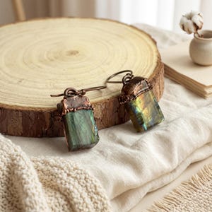 Peut inclure: Une paire de boucles d'oreilles pendantes avec des pierres de labradorite rectangulaires aux reflets irisés verts et dorés. Chaque pierre est surmontée d'un métal texturé couleur cuivre foncé et fixée à un crochet d'oreille en cuivre.