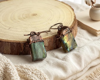 May include: A pair of dangle earrings featuring rectangular labradorite gemstones with iridescent green and gold flashes. Each stone is capped with textured, dark copper-toned metal and attached to a simple curved copper ear wire hook.
