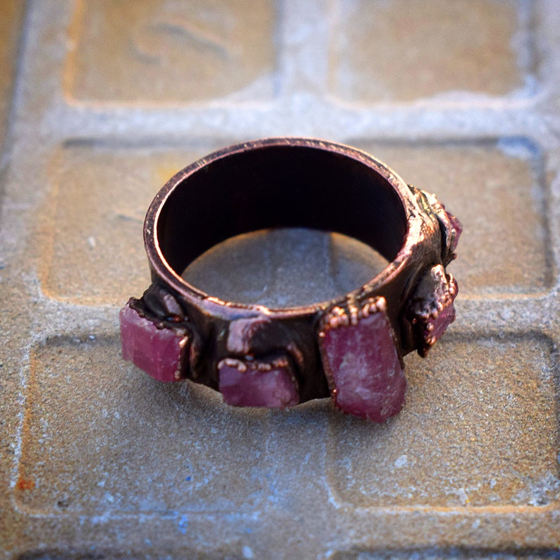 Pode incluir: Um anel de cobre com uma banda de pedras rosa e vermelhas cravadas no metal.