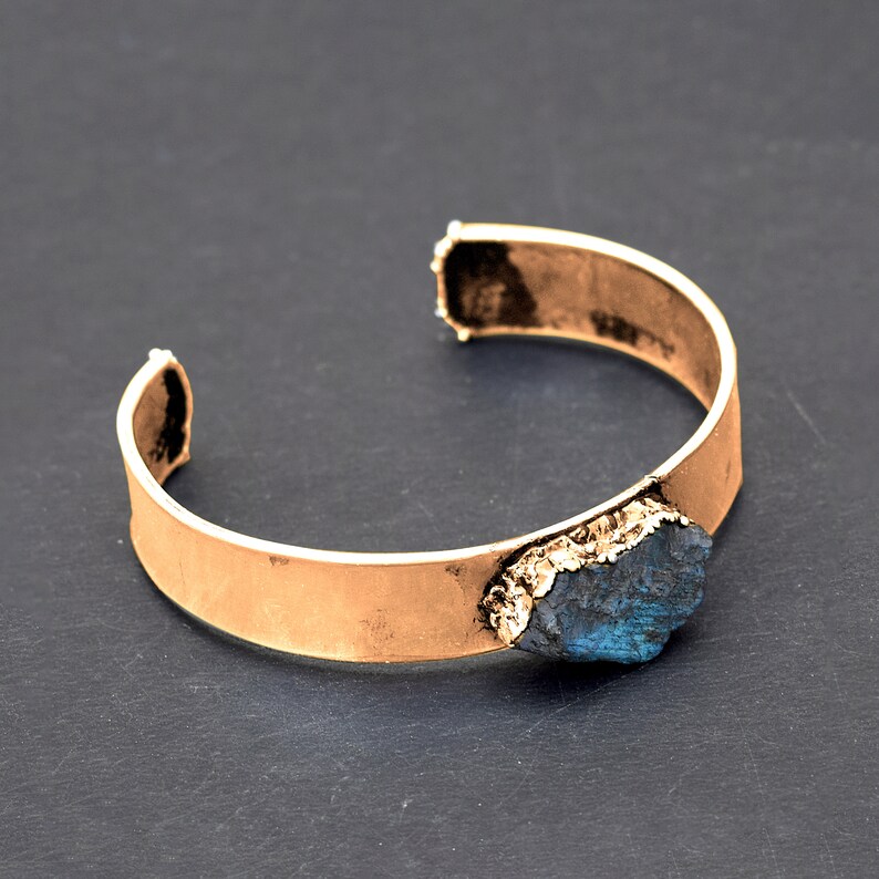 May include: A gold-toned metal cuff bracelet featuring a large, dark blue, irregularly shaped gemstone set into the metal. The gemstone has a rough, natural texture.
