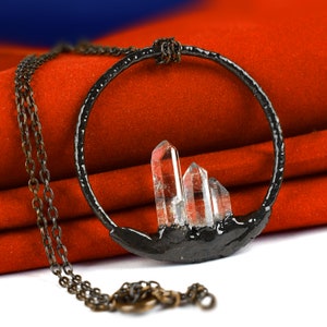 Op de afbeelding: Een zwarte metalen ketting met een ronde hanger met drie heldere kwartskristallen. De ketting ligt op een rode fluwelen achtergrond.
