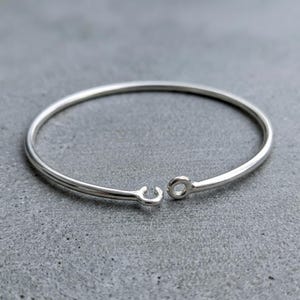 May include: A polished silver-tone metal bangle bracelet featuring a smooth, circular wire band. The bracelet has an open design with two small, rounded terminal ends, one shaped as a simple loop and the other as a small, open hook clasp.