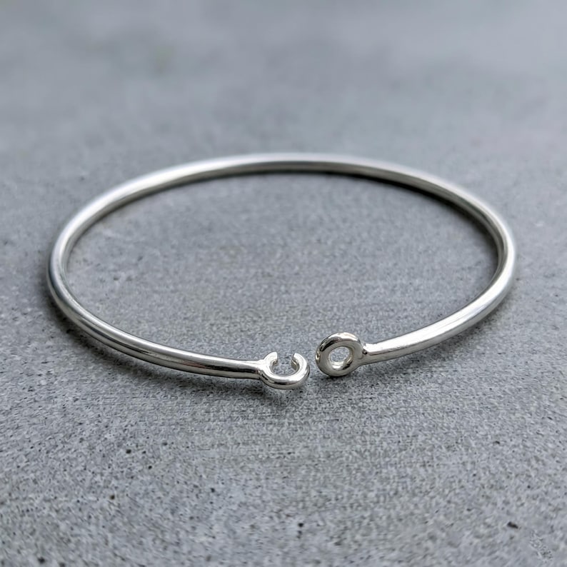May include: A polished silver-tone metal bangle bracelet featuring a smooth, circular wire band. The bracelet has an open design with two small, rounded terminal ends, one shaped as a simple loop and the other as a small, open hook clasp.