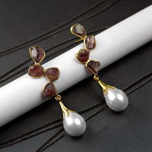 May include: A pair of gold earrings with a teardrop pearl dangle. The earrings feature a cluster of small, irregular, reddish-brown stones.