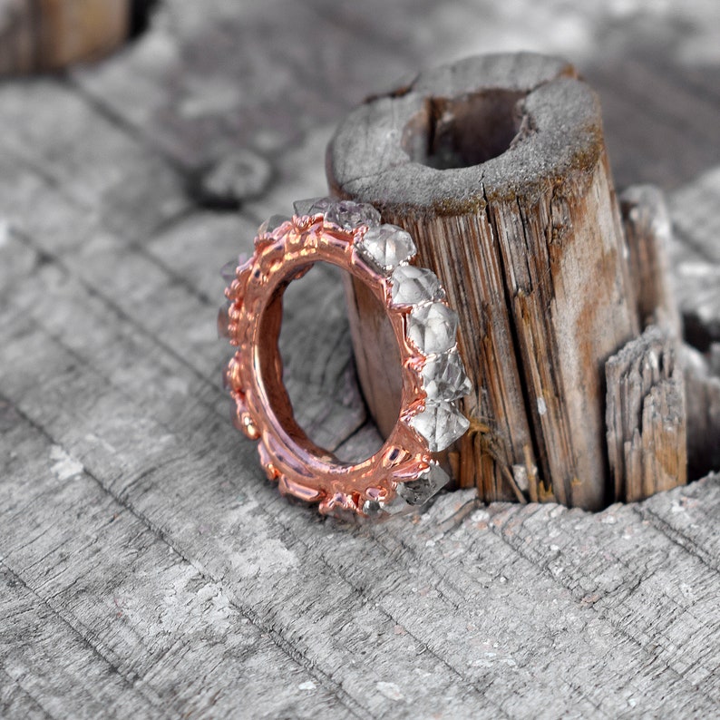 Puede incluir: Un anillo de oro rosa con una banda de peque&ntilde;as piedras facetadas transparentes. El anillo est&aacute; sobre un trozo de madera desgastada.