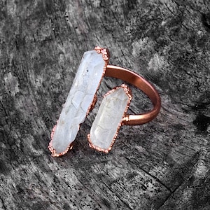 Op de afbeelding: Een koperen ring met twee witte kristallen punten. De ring is gemaakt van koperdraad en de kristallen zijn in het draad gezet. De ring ligt op een houten oppervlak.