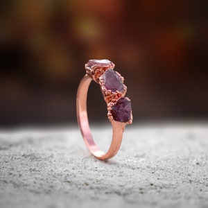 May include: A rose gold ring with three raw ruby stones set in a band. The stones are a deep red color and have a rough, natural finish.
