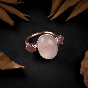 May include: A rose gold ring featuring a large, oval-shaped pink gemstone, flanked by two smaller pink gemstones.