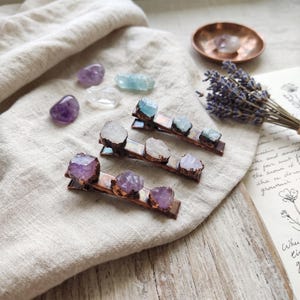 May include: Three metal hair clips featuring raw, unpolished crystal stones. One clip has light blue stones, one has clear white stones, and one has purple amethyst stones. The metal base of each clip has a dark, oxidised copper-toned finish.
