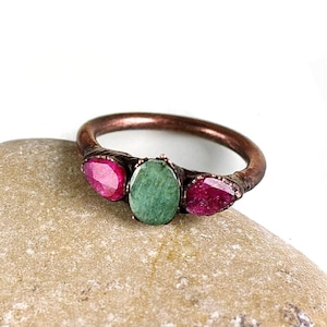 May include: A copper ring with three gemstones: a green emerald in the center and two pink rubies on either side. The ring is set on a rough, brown stone.